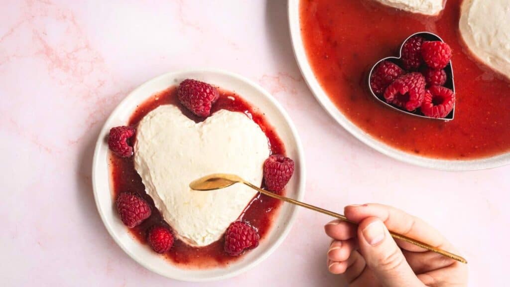 A person holding a spoon and a heart shaped dessert with raspberries.