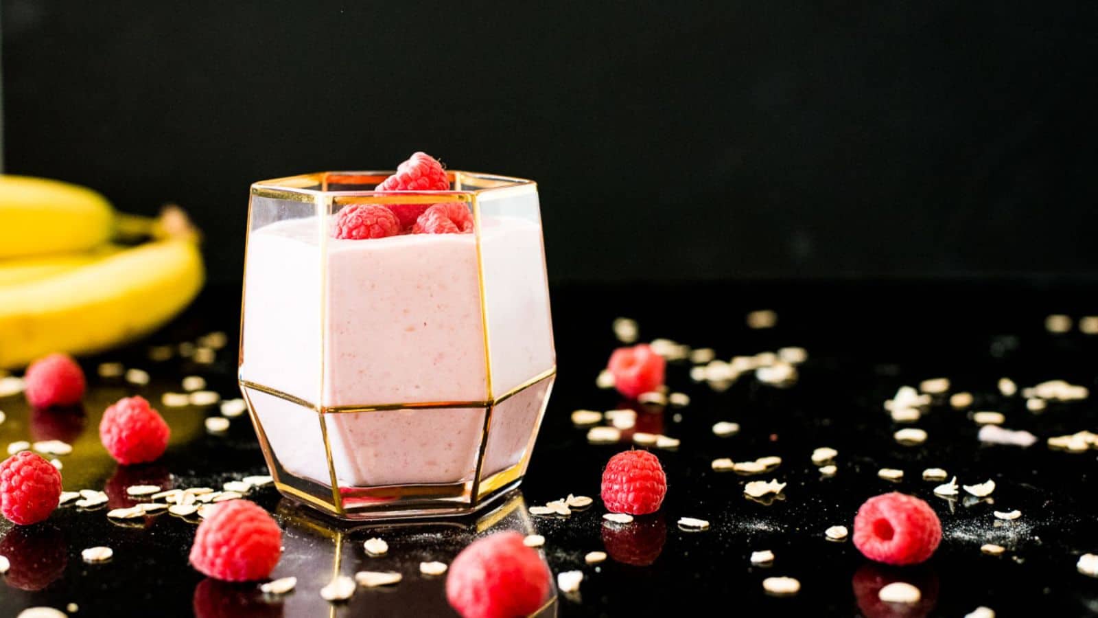 Raspberry smoothie with raspberries on top in gold-trimmed glass on a black surface.