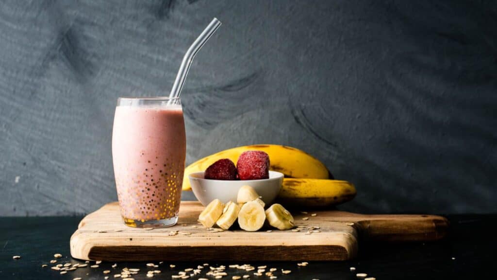 A strawberry banana oat smoothie with glass straw in front of strawberries and bananas on a wooden board, on a dark blue background with text overlay.