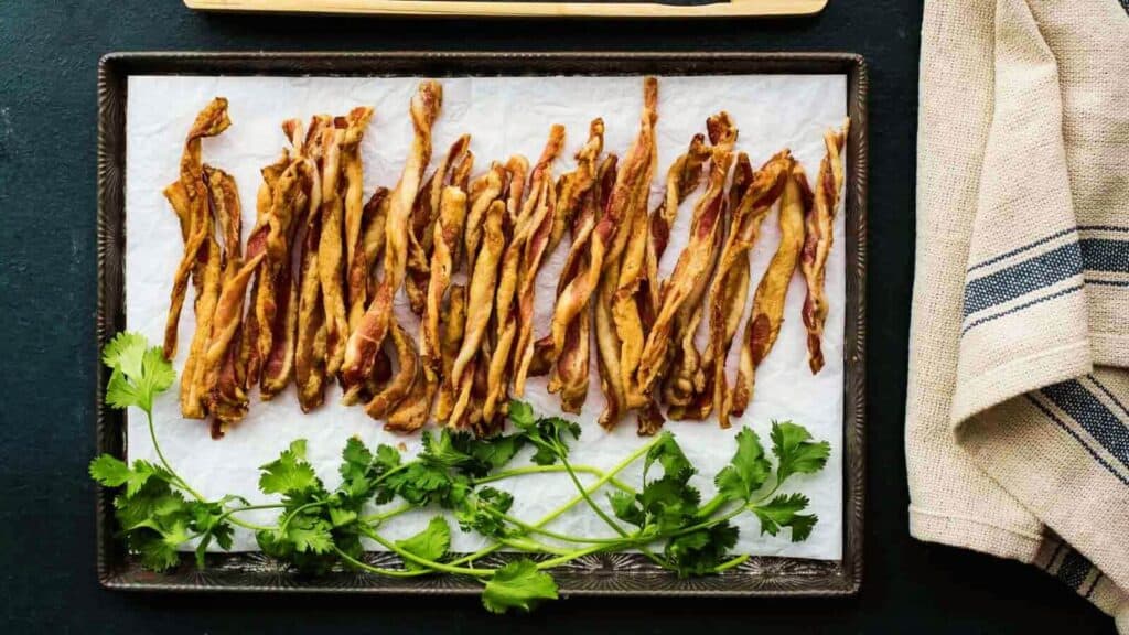 A baking sheet with twisted bacon sticks and parsley on it.