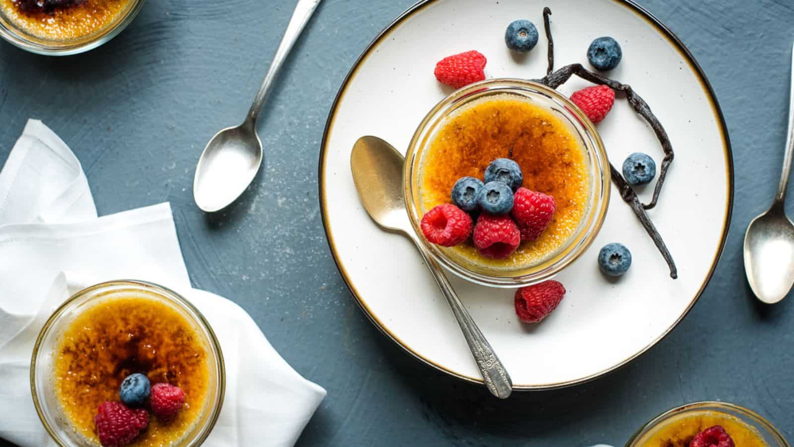 Overhead view of three servings of vanilla creme brulee on white plates and napkins with a blue background.