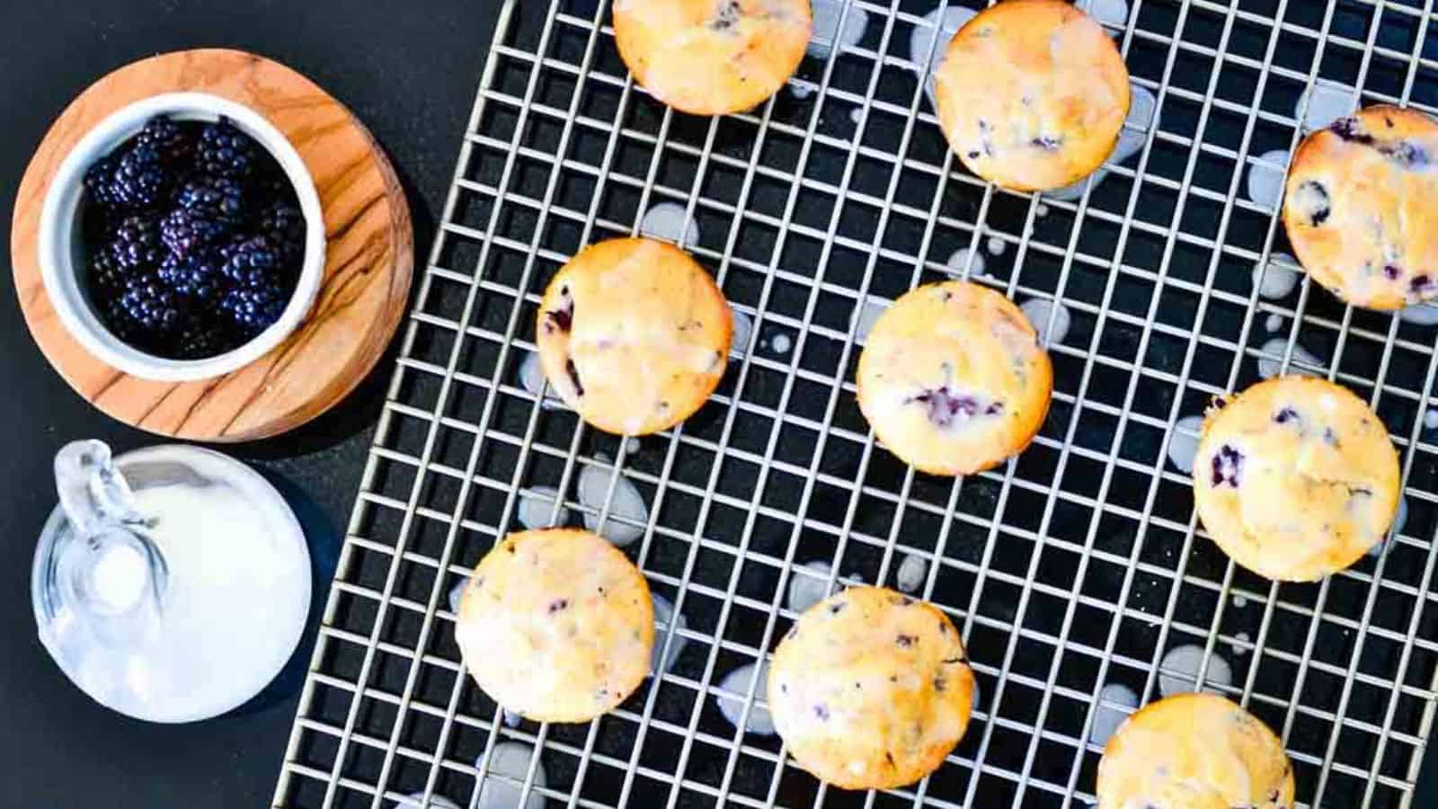Blackberry muffins drizzled with lemon glaze sitting on a gray cooling rack on a dark background.