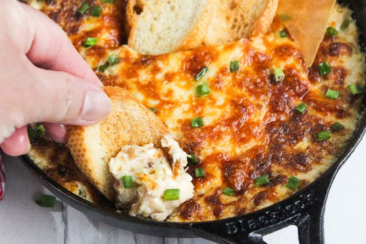 A person dipping a piece of bread into a skillet of cheesy dip.