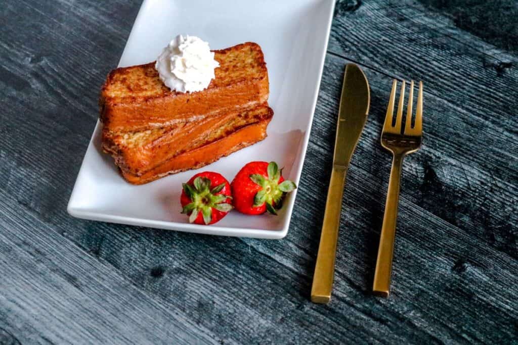 A sweet slice of french toast with luscious strawberries and a dollop of whipped cream on a white plate.