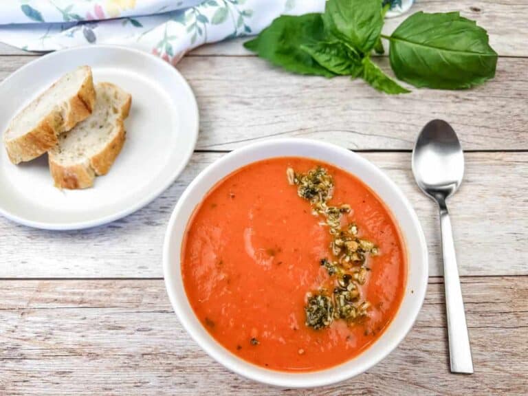 A bowl of red pepper and tomato soup next to a plate of bread and a spoon.