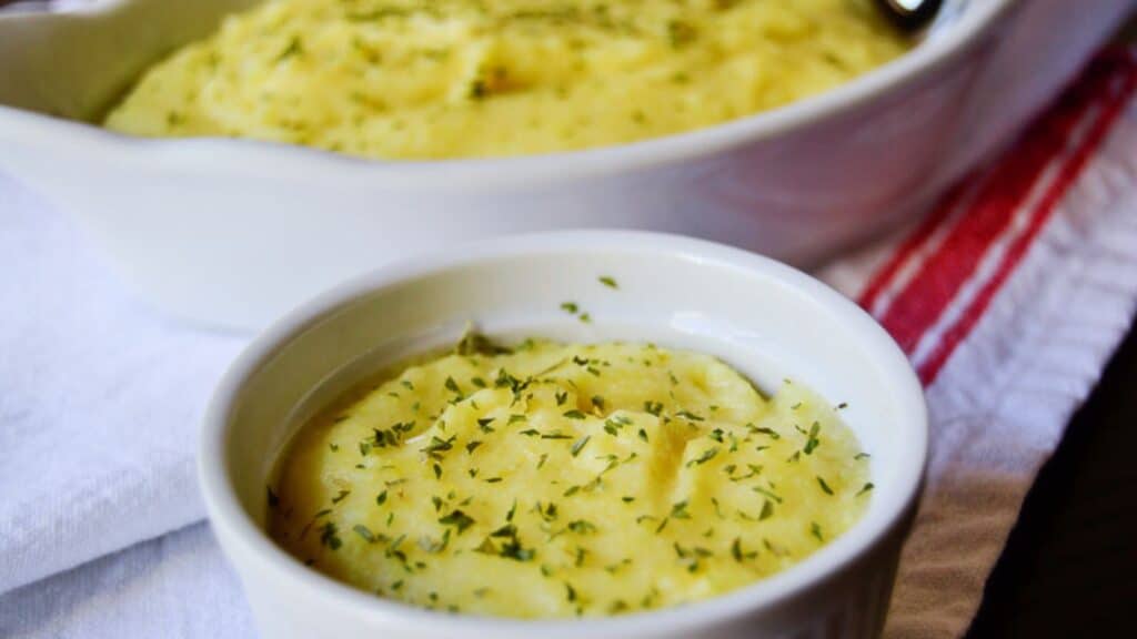 Baked cheesy mashed potatoes garnished with herbs in a white ramekin and a larger dish in the background.