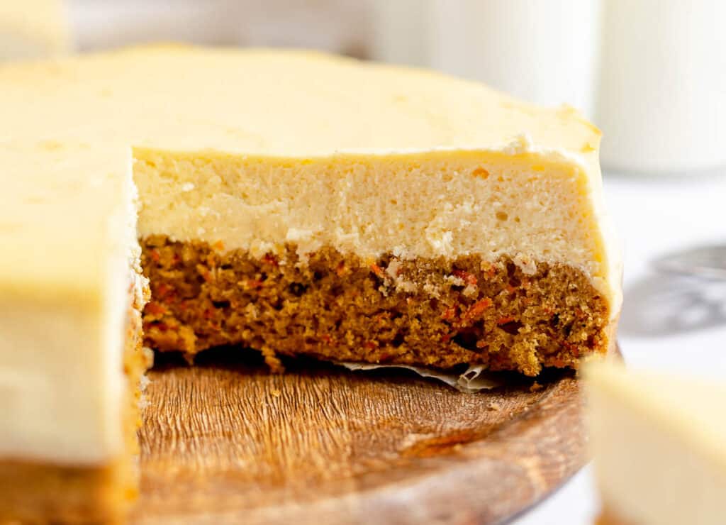 A slice of carrot cheesecake on a wooden plate with a creamy layer on top.