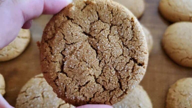 A close-up of a hand holding a freshly baked ginger snap cookie with more cookies in the background.