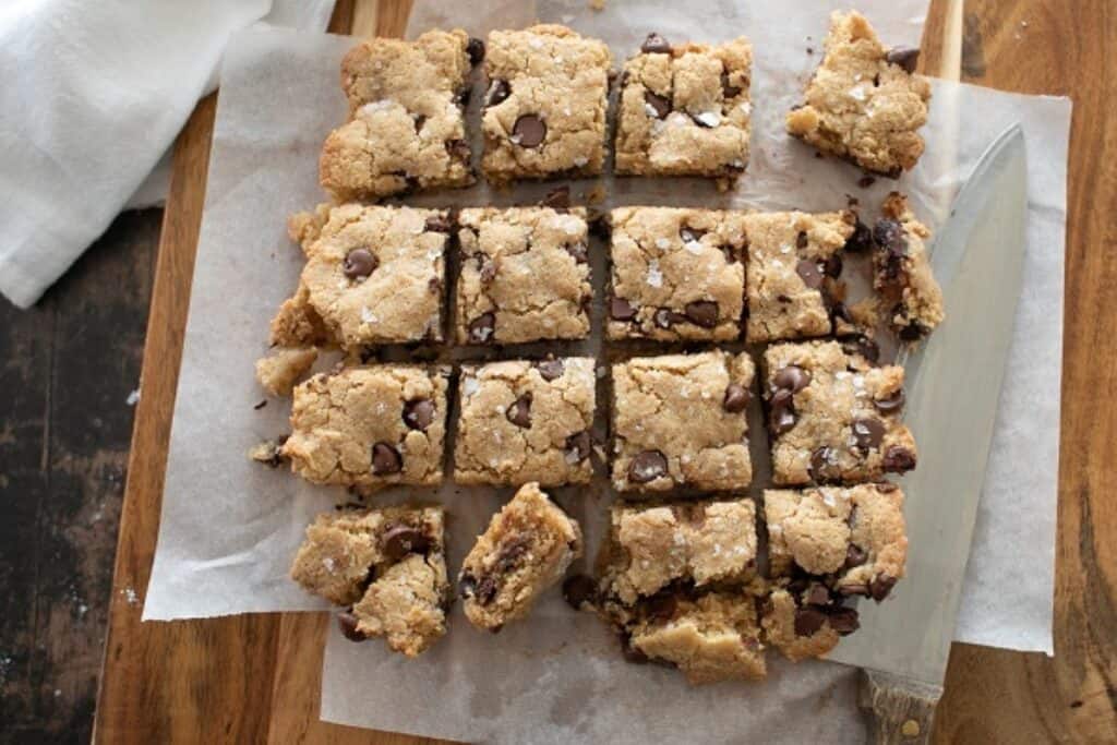 A parchment paper-lined tray with freshly baked chocolate chip blondies cut into squares, accompanied by a knife.