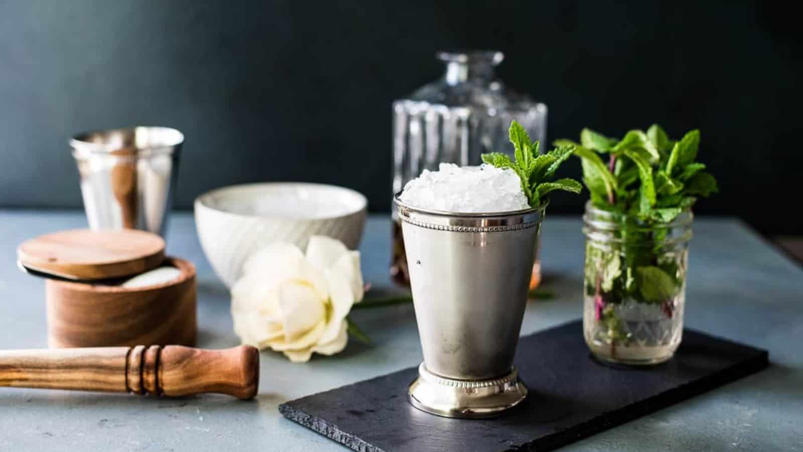A mint julep in a silver mint julep cup with sprigs of mint and a decanter of bourbon and other ingredients in the background.