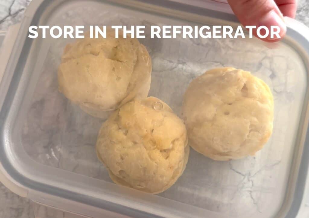 A person is holding two biscuits in a container with the words store in the refrigerator.