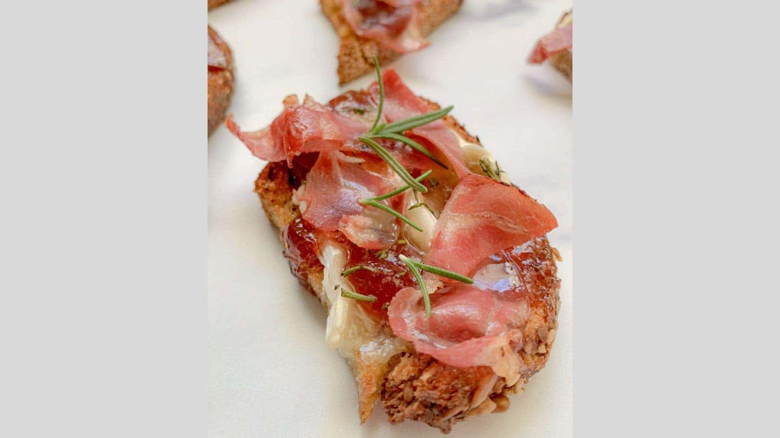 A slice of crisp bread spread with brie cheese, crispy prosciutto, and raspberry preserves.