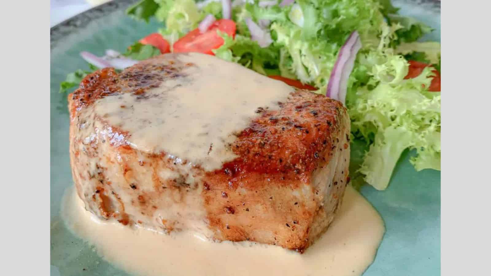 A thick cut pork chop on a plate drizzled with a mustard au jus and a small salad.