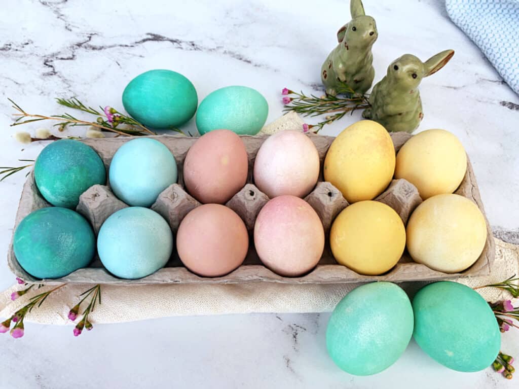 A carton of pastel-colored easter eggs with two decorative bunny figures next to blooming flowers.