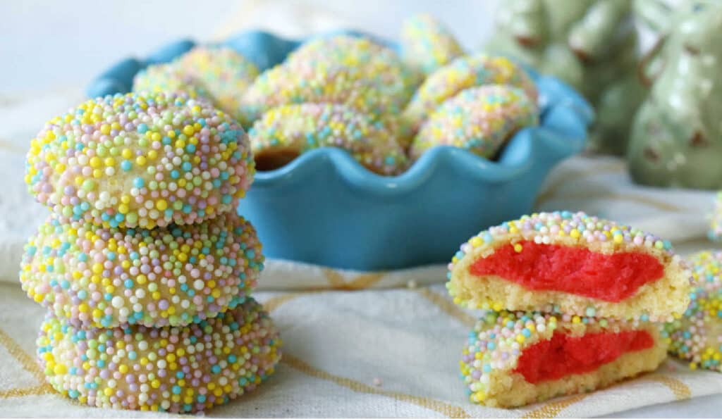 Colorful sprinkle-coated cookies, some whole and one bisected revealing a red interior, presented in a blue dish.