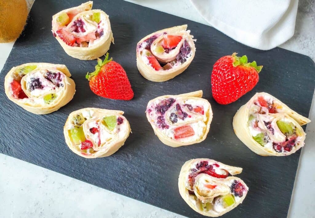 Assorted fruit and cream cheese pinwheel sandwiches on a slate board with fresh strawberries.