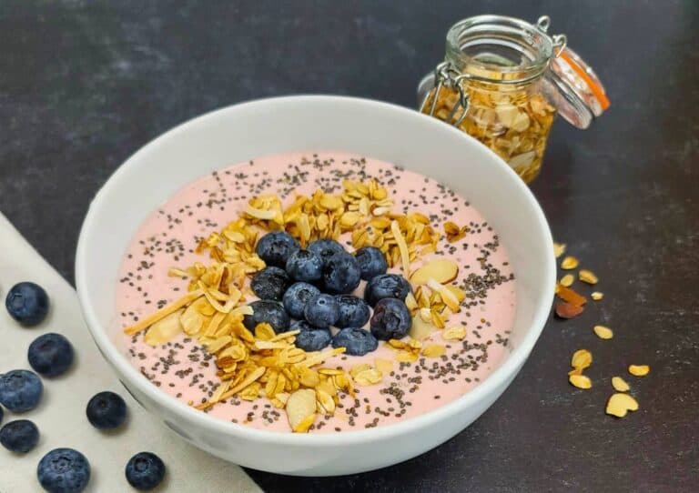 A bowl of pink smoothie topped with granola, blueberries, and chia seeds, accompanied by a jar of granola and scattered blueberries on a dark surface.