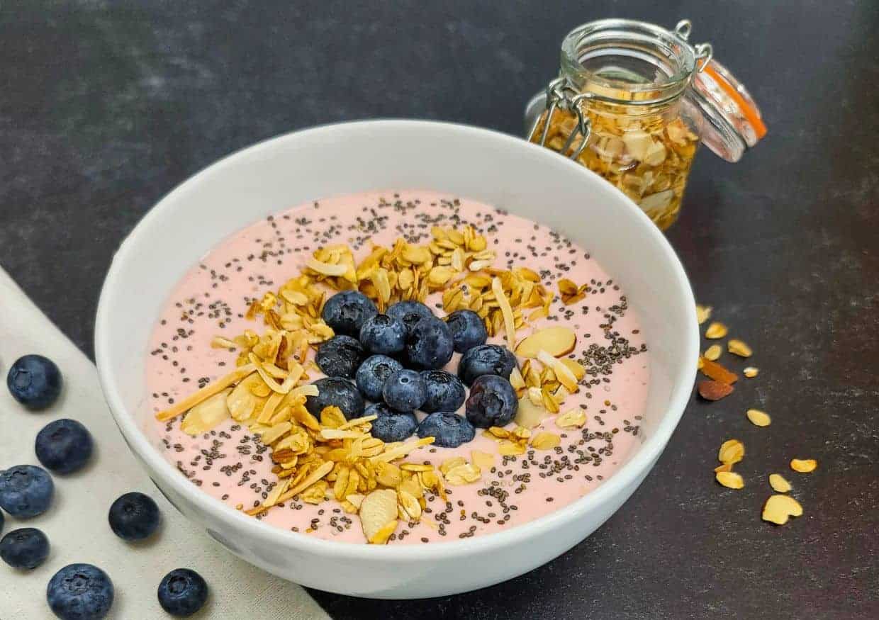 A bowl of pink yogurt topped with granola, chia seeds, and fresh blueberries, with extra blueberries and a jar of granola on the side.