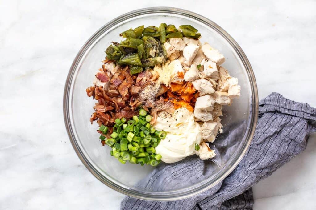 A bowl containing ingredients for a recipe, including chopped green onions, bacon bits, shredded chicken, cheese, and spices.