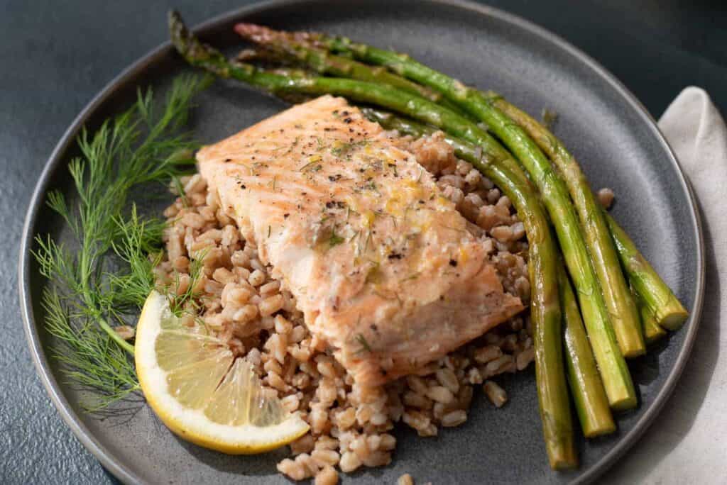A serving of lemon dill salmon and asparagus served over faro, garnished with dill and a lemon slice.
