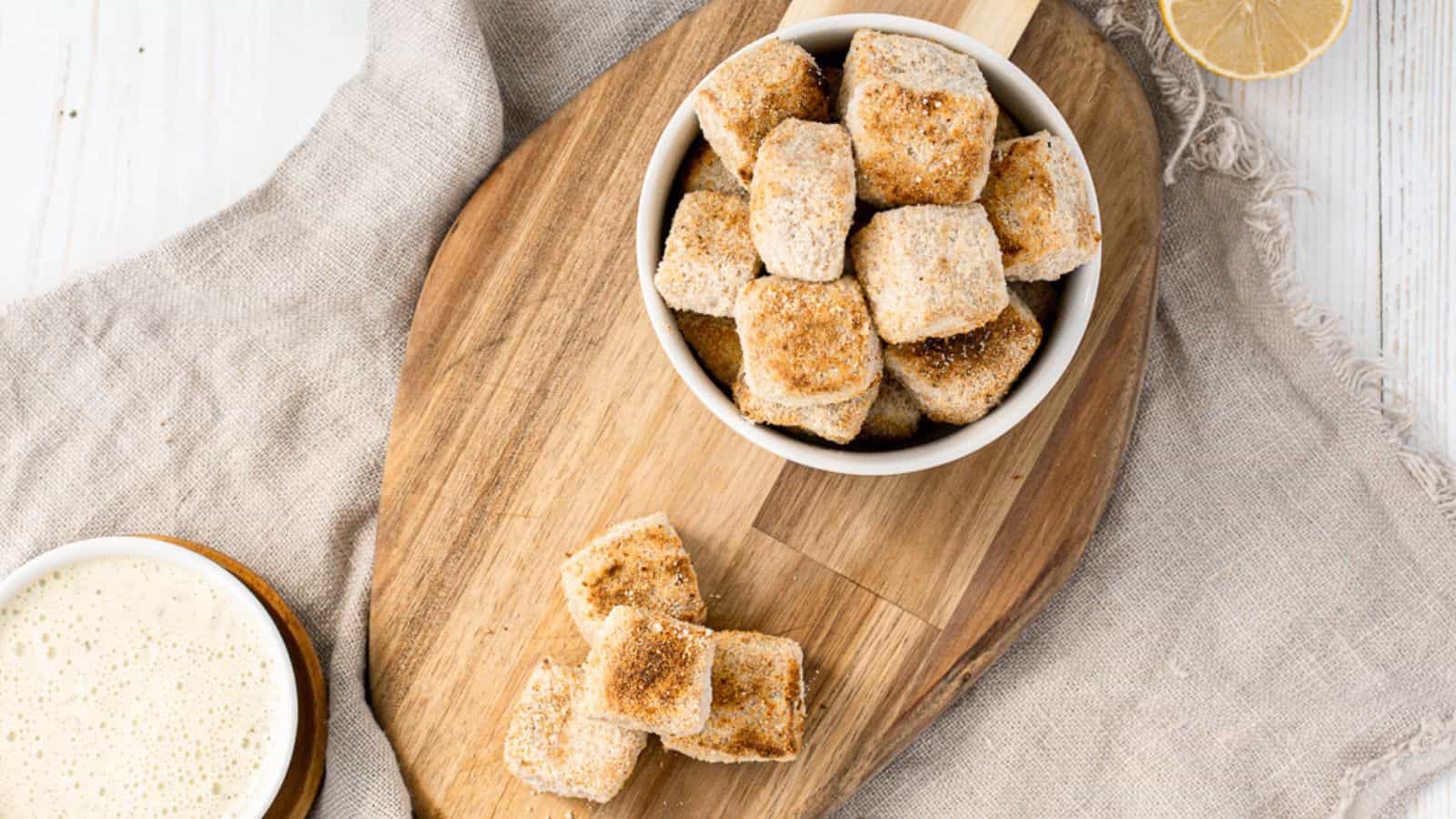 A wooden cutting board with a bowl of tofu bites and a bowl of dipping sauce.