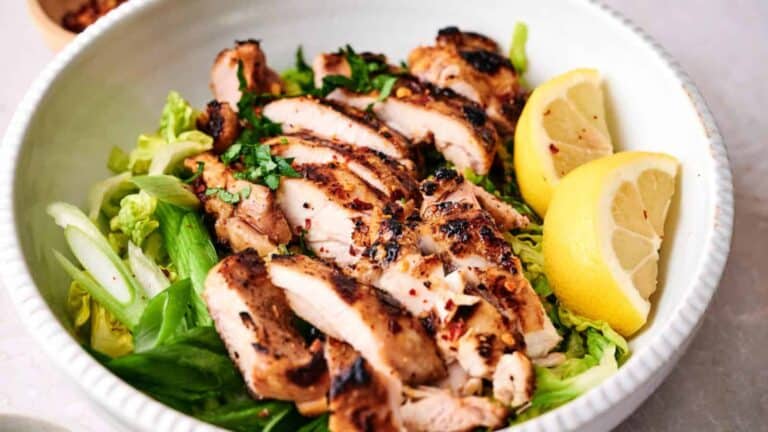 Grilled chicken breast slices on a bed of salad greens with lemon wedges on the side.
