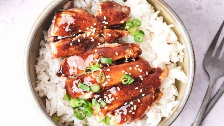 A bowl of white rice topped with glazed chicken and garnished with green onions and sesame seeds.