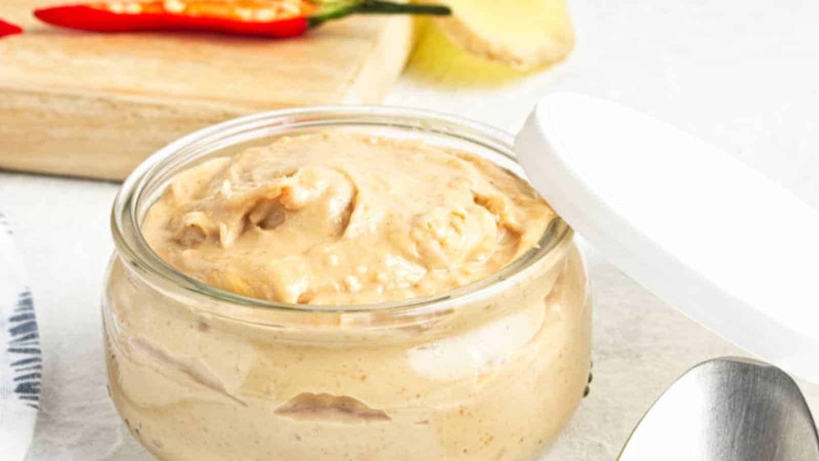 A jar of creamy peanut sauce with a spoon on the side, flanked by fresh peppers and peanuts.