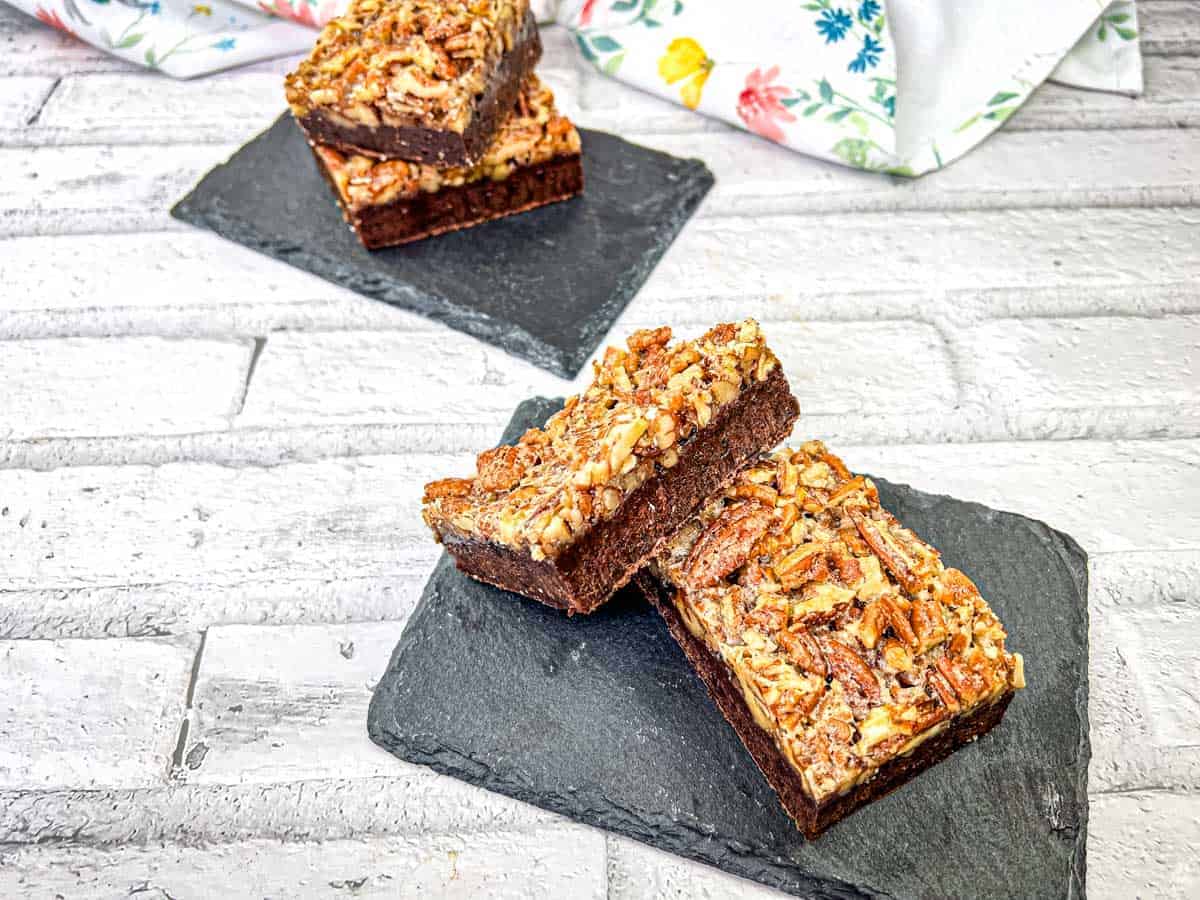 A close-up of two pecan brownies on a slate surface, showing the rich, chocolatey interior and a topping of chopped pecans.