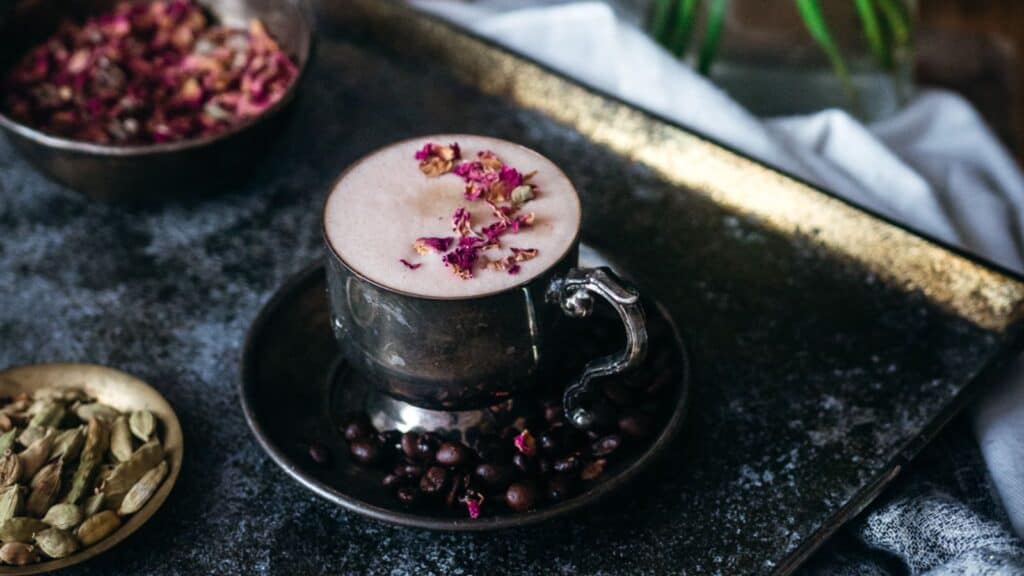 A cup of coffee garnished with rose petals, resting on a saucer surrounded by coffee beans, with cardamom pods on a plate and a rustic background.