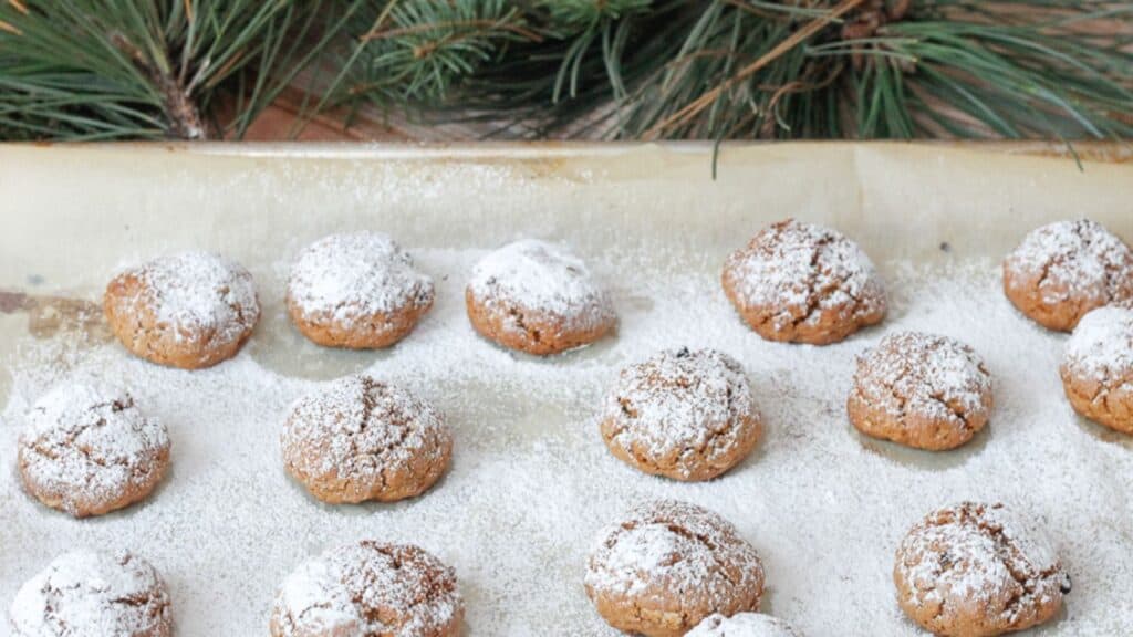 Freshly baked cookies dusted with powdered sugar on parchment paper, with pine branches in the background.