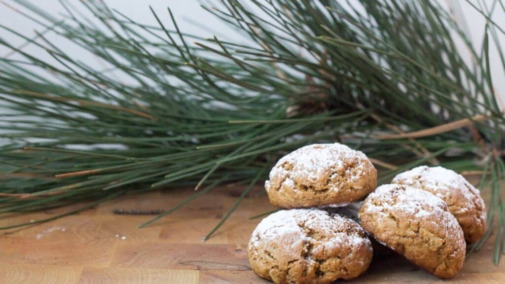 Fresh-baked cookies dusted with powdered sugar beside pine branches on a wooden surface.