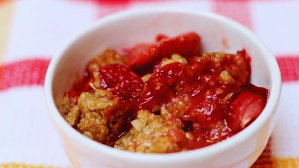 A bowl of strawberry rhubarb crisp on a red and white striped cloth.