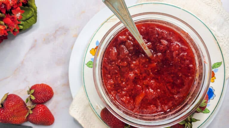 A jar of homemade strawberry jam on a plate with a spoon inside, accompanied by fresh strawberries on a marble surface.