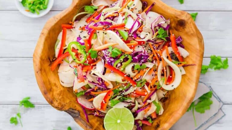 A colorful mixed vegetable slaw with red cabbage, carrots, and herbs in a wooden bowl, garnished with lime.