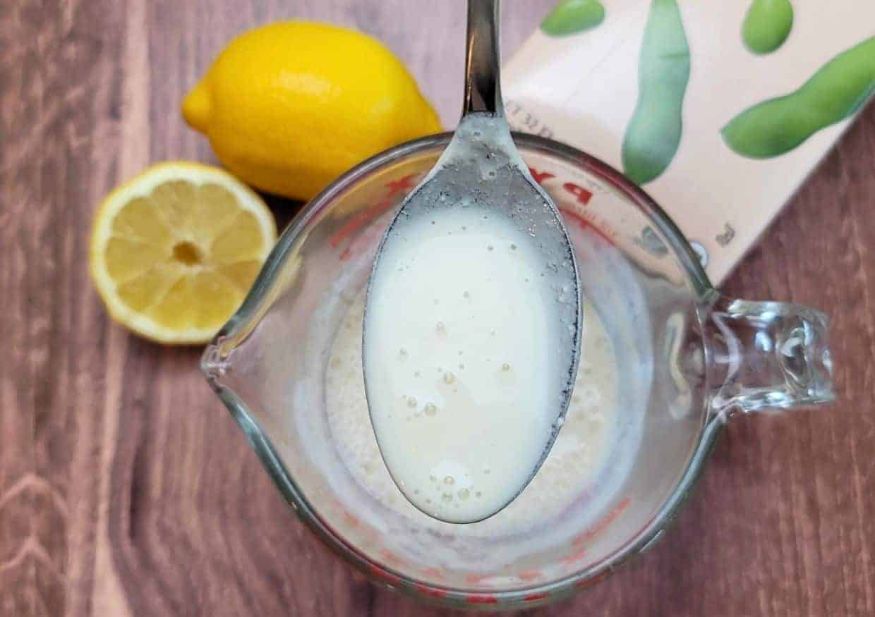 A measuring cup with a vegan buttermilk on a spoon above it, with cut lemons and a plant-based milk carton in the background.
