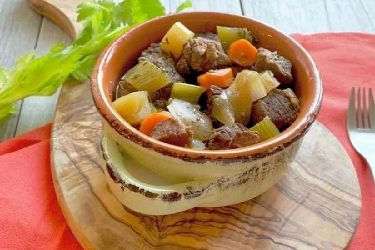 A bowl of stew with meat and vegetables on a wooden cutting board.