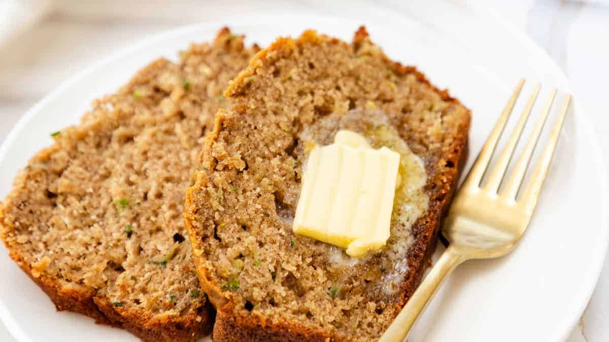 The image displays slices of moist zucchini bread with butter on a plate, alongside a fork.