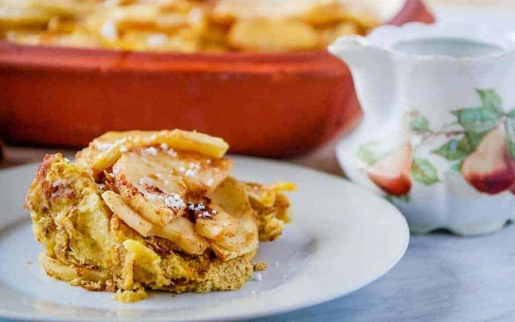 Piece of French toast casserole on a plate with a dusting of powdered sugar and a pitcher of syrup.