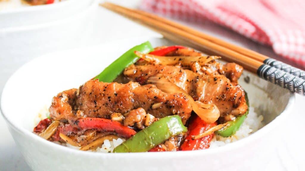 A bowl of chicken stir-fry with vegetables served over white rice with chopsticks on the side.