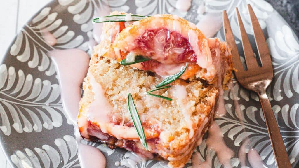 A slice of lemon raspberry loaf with a drizzle of glaze, garnished with a sprig of rosemary, on a patterned plate with a fork beside it.