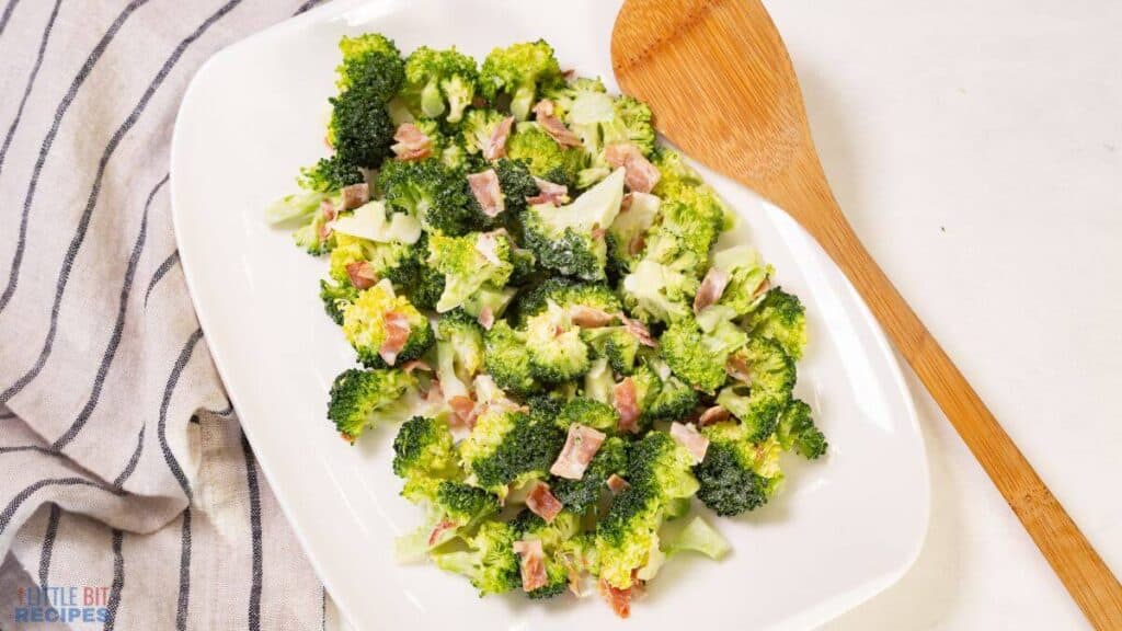 A dish of broccoli salad with bacon and cheese, served on a white plate with a wooden spoon on the side.