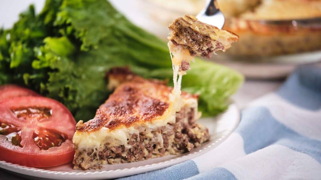 A slice of meat pie with melted cheese alongside fresh tomato slices and lettuce.