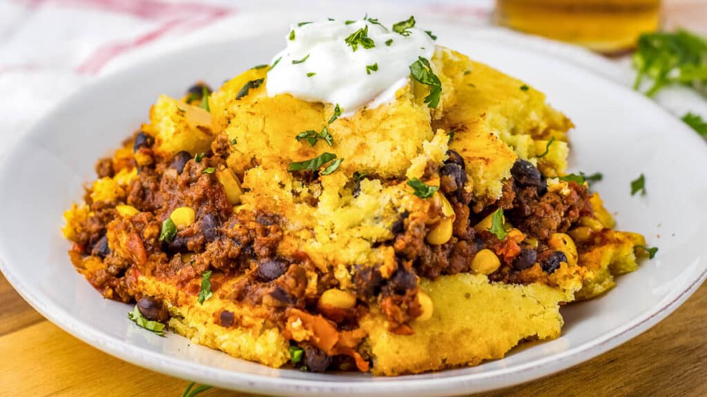 A plate of cornbread topped with chili con carne and a dollop of sour cream, garnished with chopped herbs.