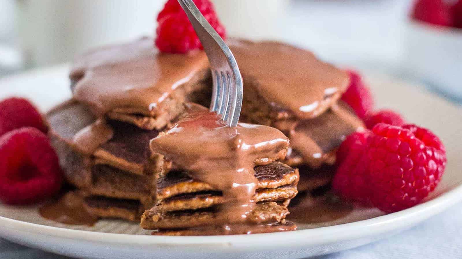 Stack of chocolate protein pancakes with chocolate sauce and raspberries on a plate.