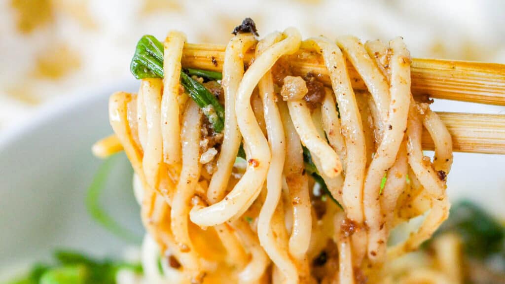 Chopsticks holding a twirl of spaghetti with green onions and visible seasoning.