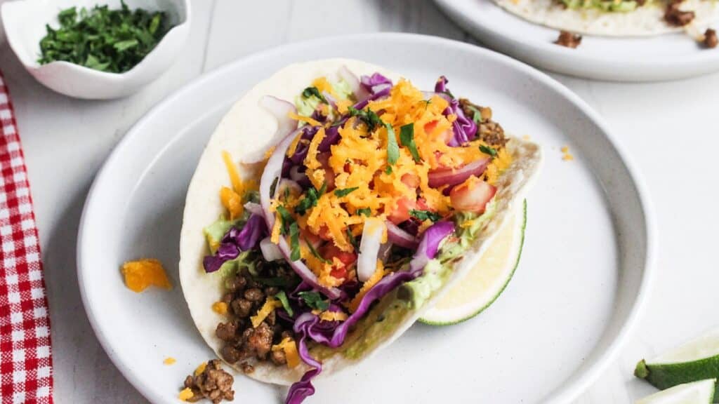 A colorful taco filled with ground meat, lettuce, shredded purple cabbage, carrots, and a slice of lime on the side.