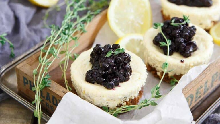 Mini cheesecakes topped with blueberry compote, garnished with fresh thyme, presented on a wooden tray.