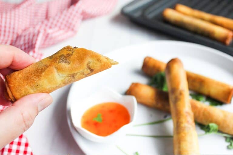 A person holding a spring roll with more rolls and dipping sauce in the background.