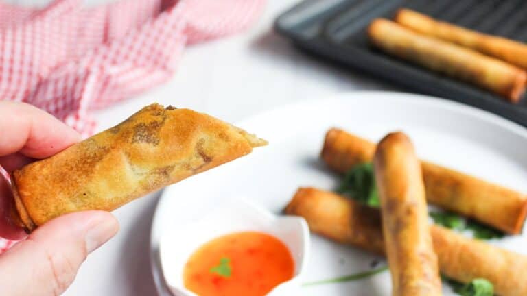 A person holding a spring roll with more rolls and a dipping sauce in the background.